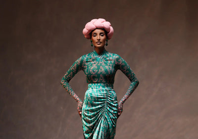 Woman in a green patterned dress with a pink headpiece against a brown background